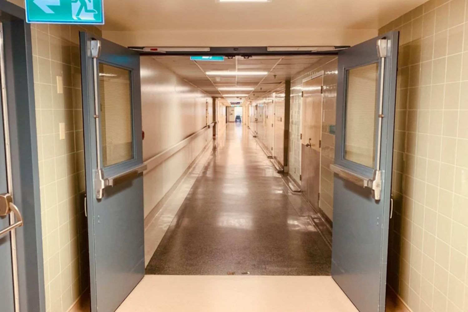 Perspective of a long illuminated hospital corridor, framed through glass fire doors with panic bars